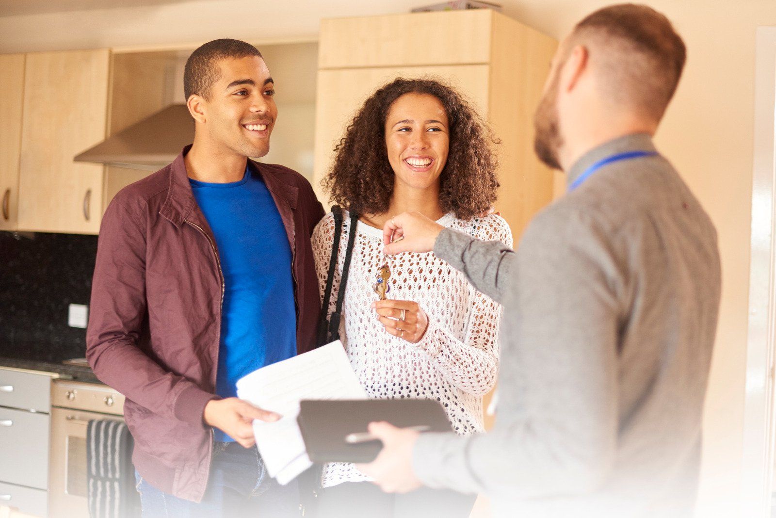 A real estate agent hands keys to two smiling people inside a residential kitchen.