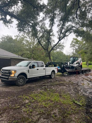 White pickup truck and equipment trailer on a muddy driveway beside a house and trees.