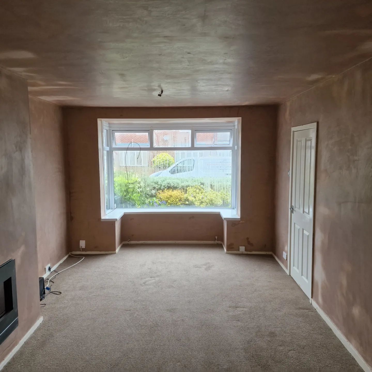 Empty room with new plaster walls, large window, carpet, and a closed door.