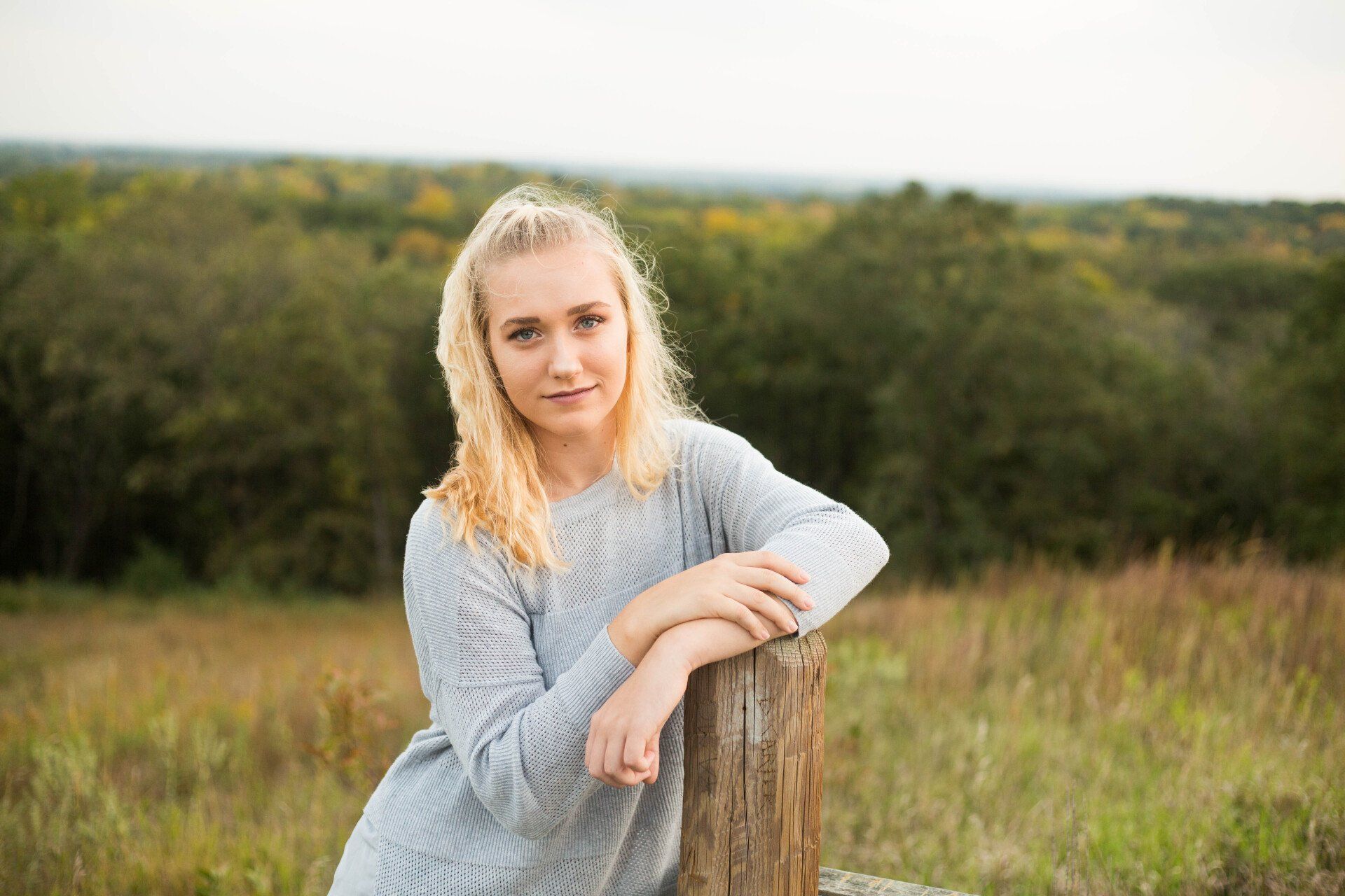 Blonde woman in gray sweater leans on a wooden post, smiling outdoors, fields and trees in the background.