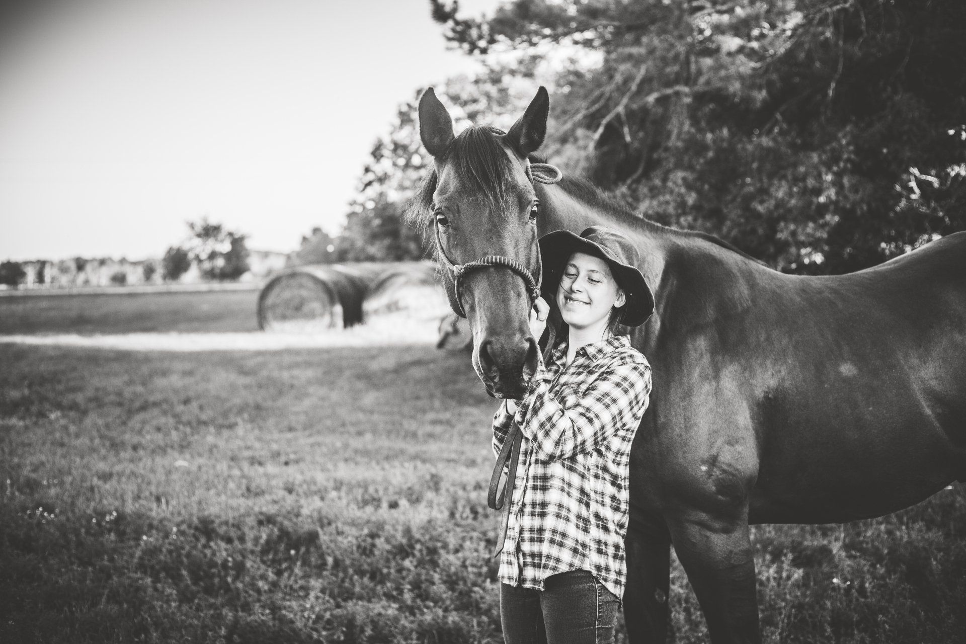 Girl in a plaid shirt smiles, petting a brown horse in a field.