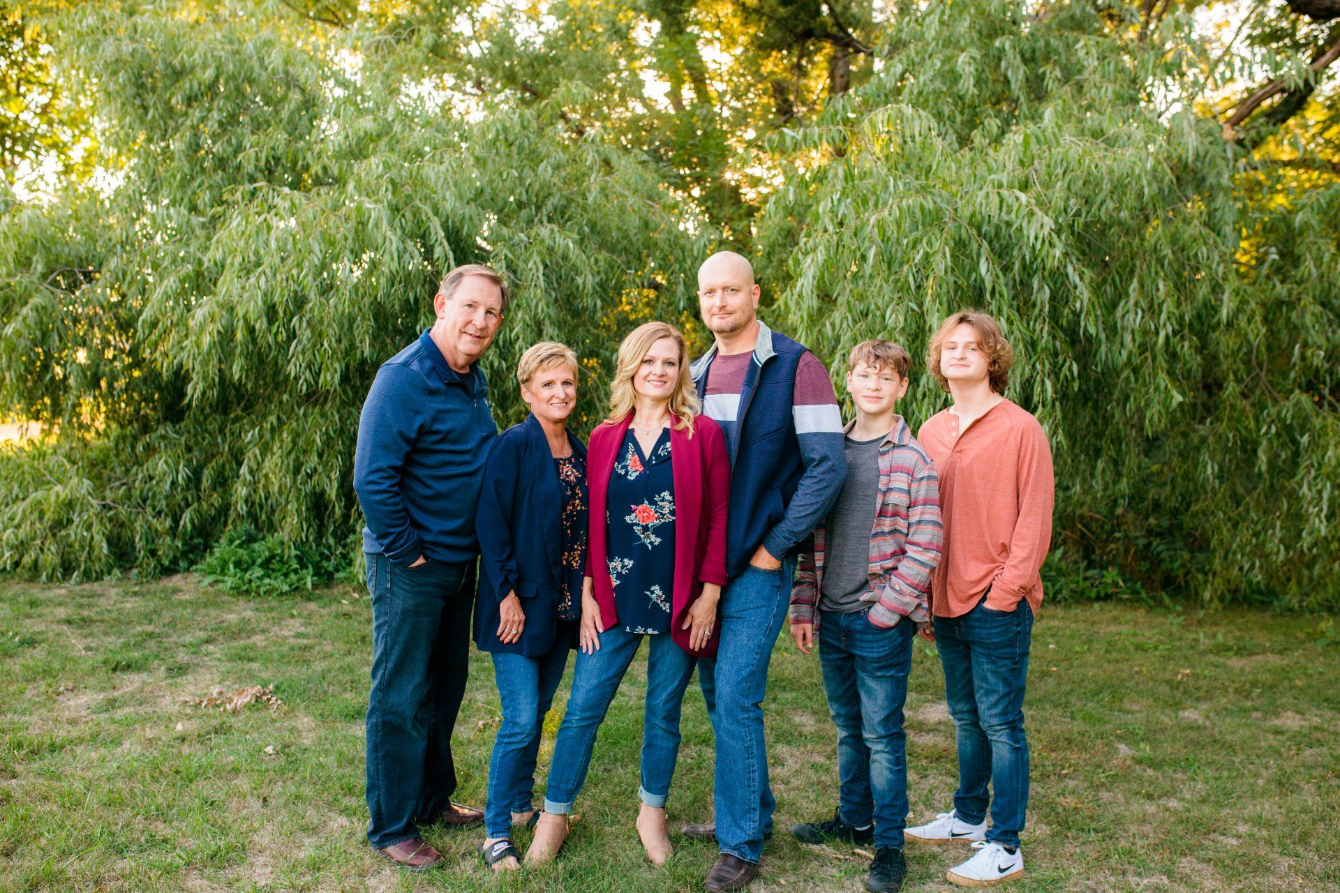 Family of six posing outdoors in front of trees; smiling, dressed in various casual outfits.