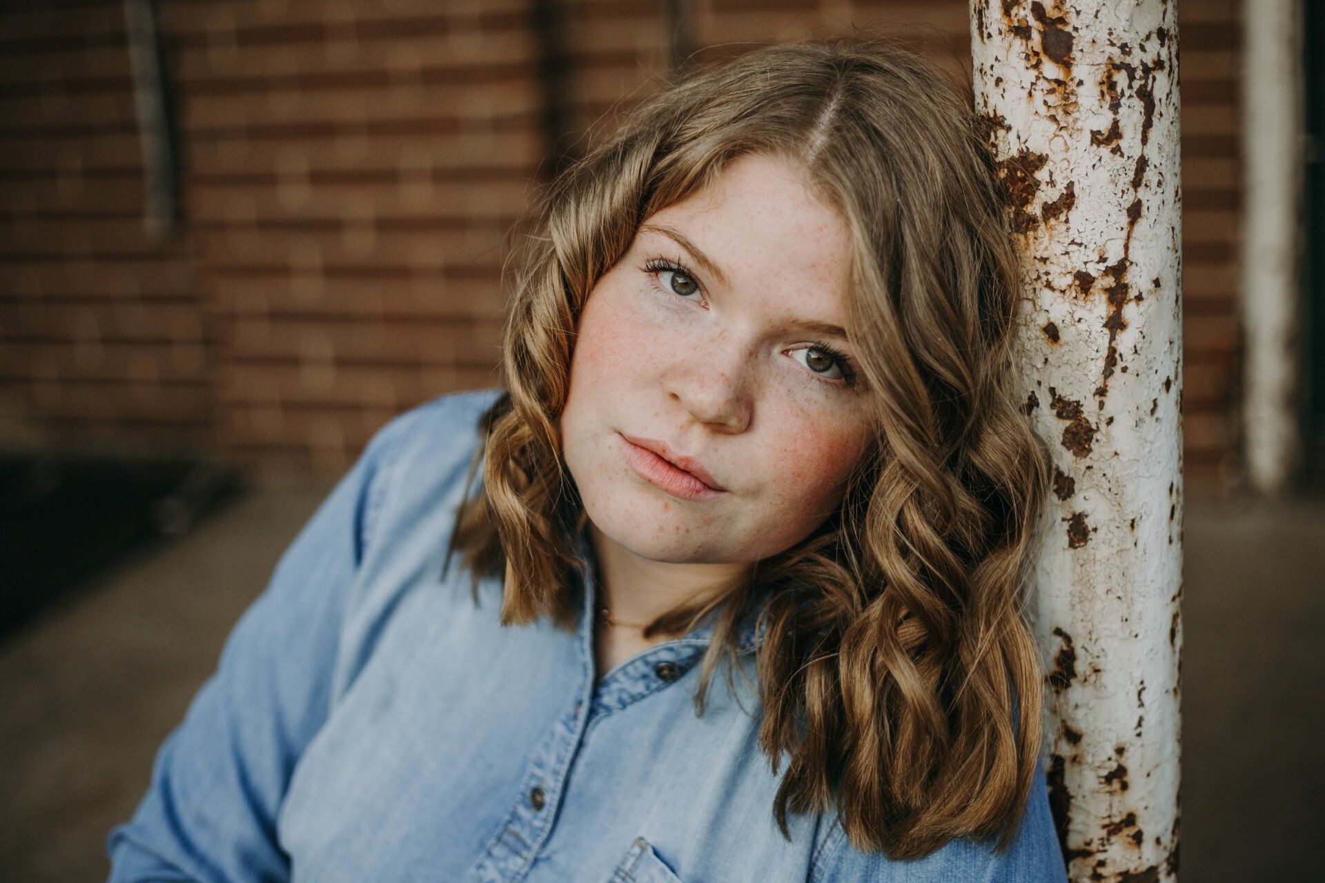 Young woman with curly blonde hair, leaning on a rusty pole, wearing a denim shirt, brick background.