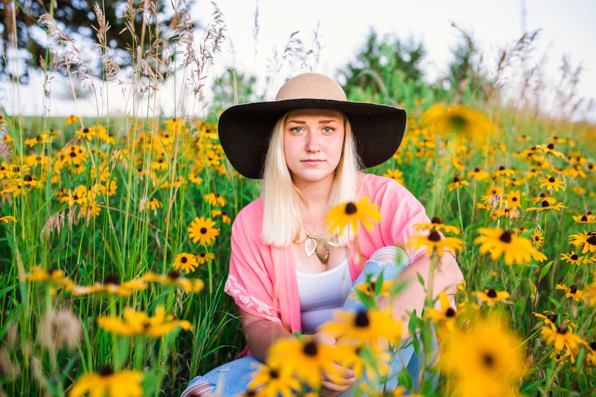 Blond woman in sun hat sits among yellow flowers, wearing pink shawl and blue patterned pants.