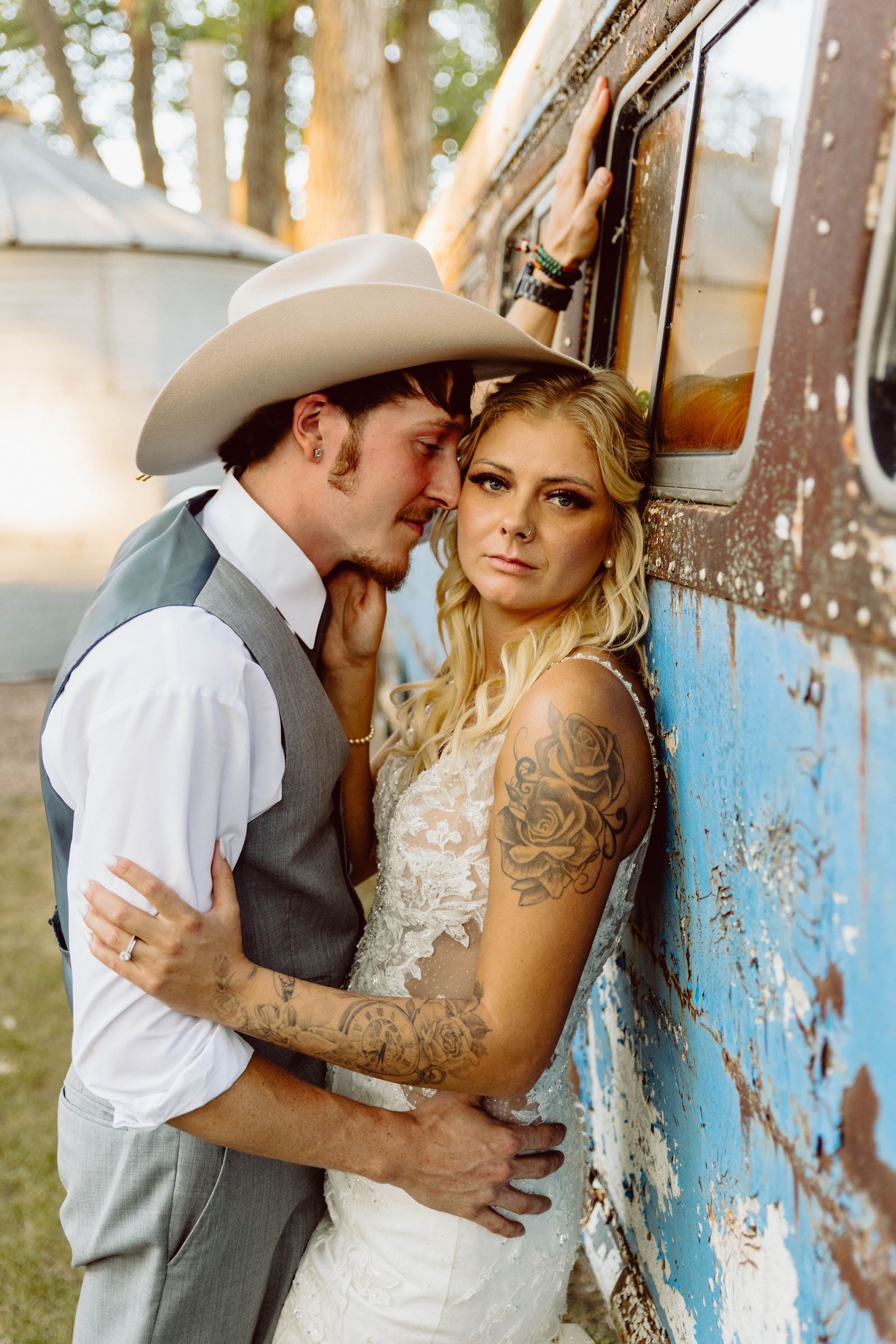 A couple in wedding attire, leaning against a weathered blue wall. Groom in cowboy hat whispers to bride.