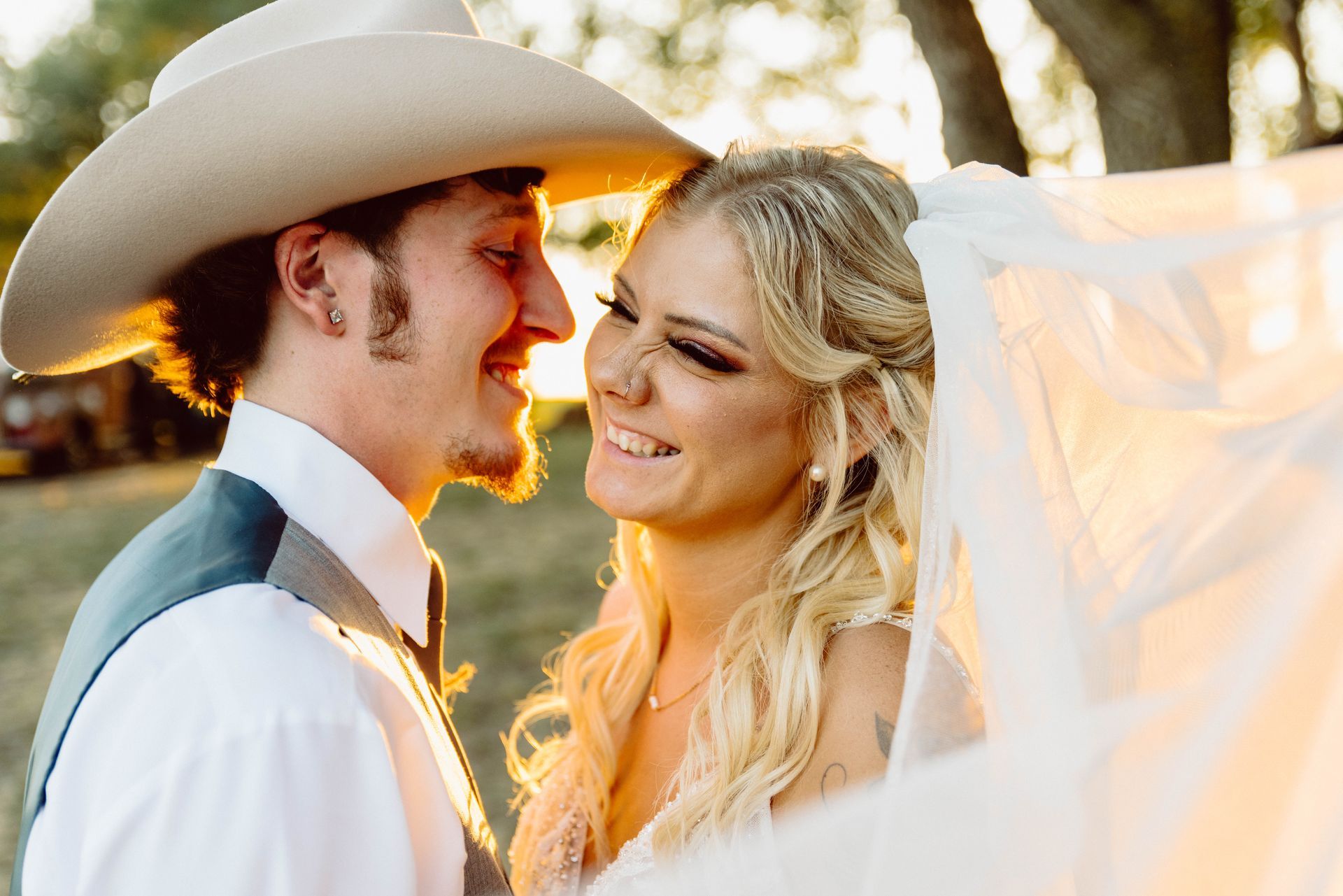 Bride and groom smiling at each other during a wedding. The groom is wearing a cowboy hat. Soft sunlight.