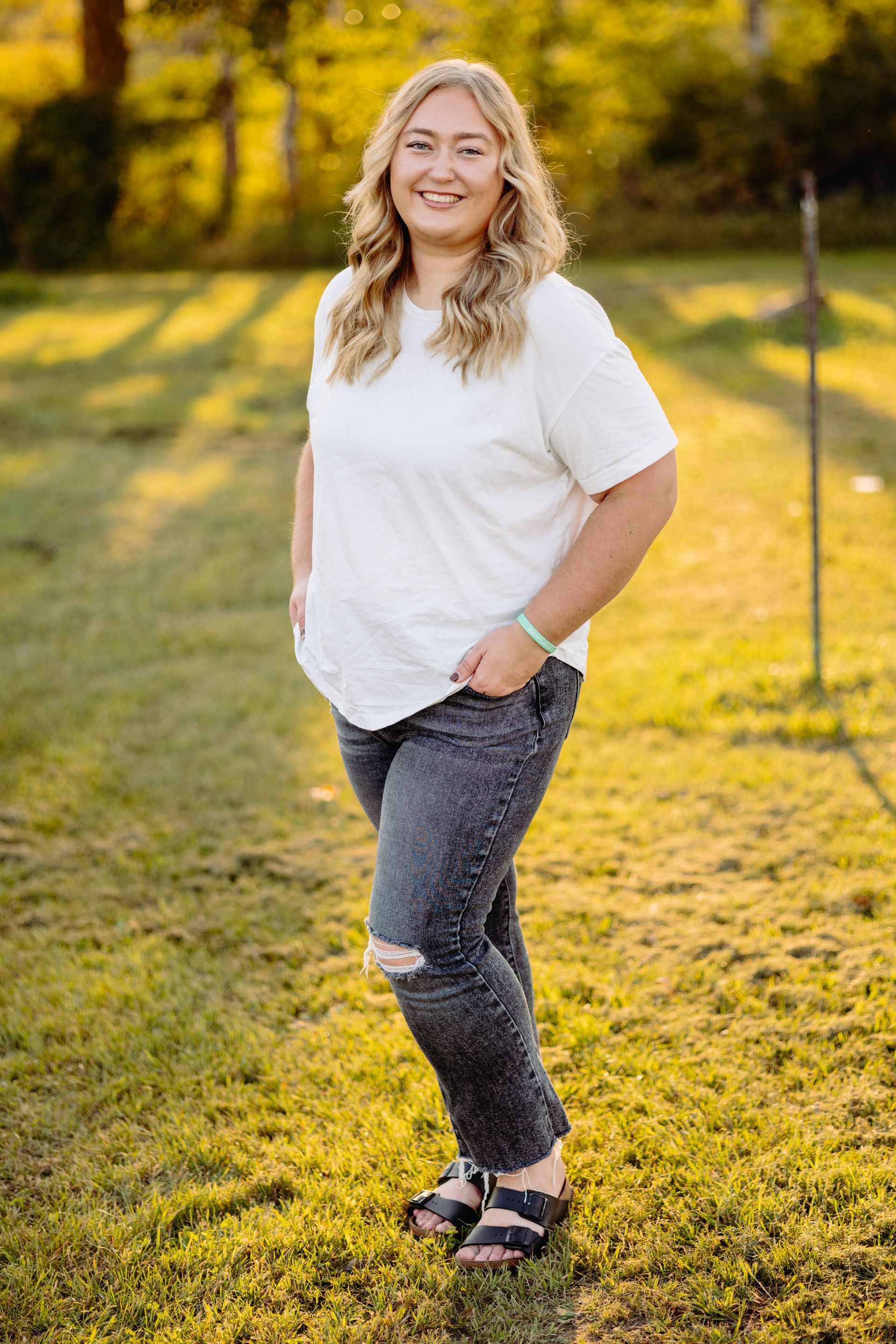 Woman with blonde hair smiling, wearing a white shirt, patterned jeans, and sandals, standing in a grassy field.