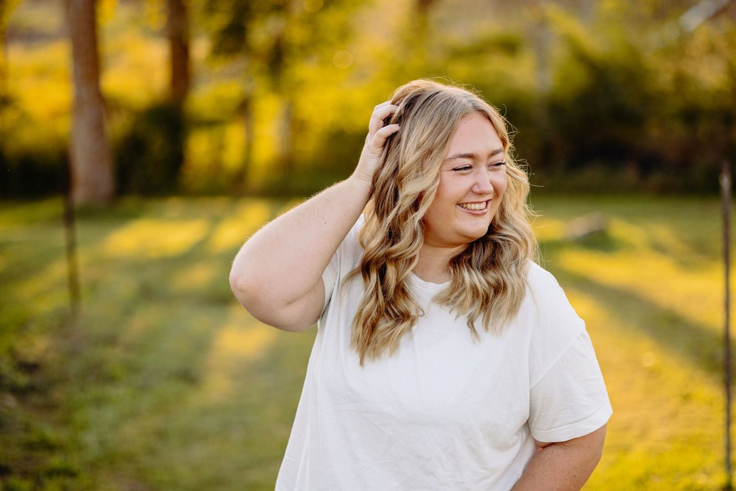 Woman with blonde hair smiling and touching her hair in a sunlit outdoor field.