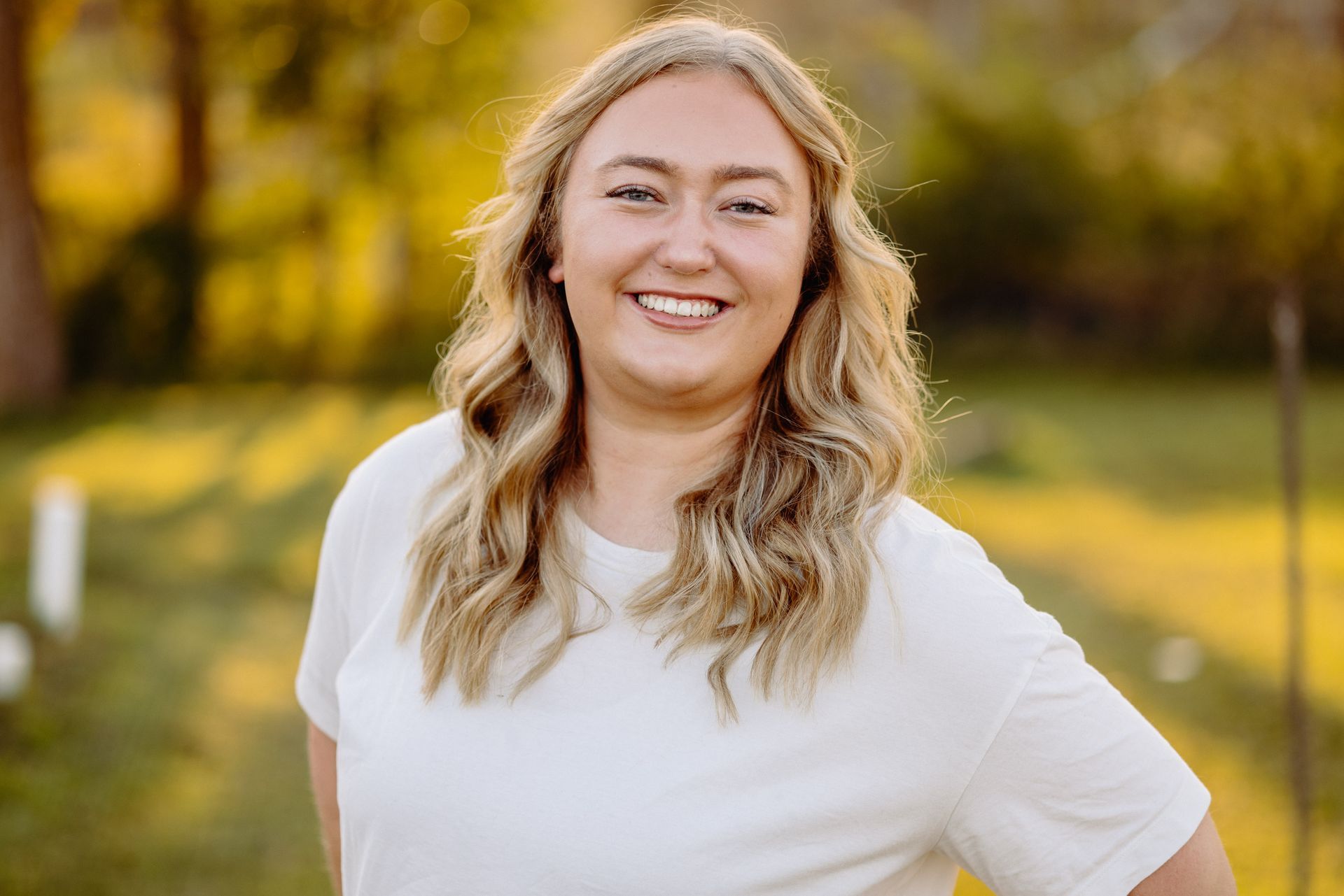Blonde woman smiling outdoors, wearing a white shirt with soft, natural lighting.