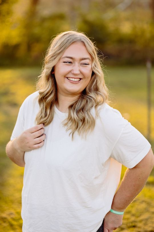 Woman who's a Minnesota portrait photographer with long blonde hair, smiles in a field with a hand on her chest.