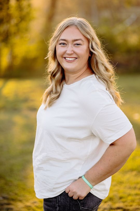 Woman, MN Photographer, with wavy blonde hair smiles, wearing a white t-shirt, and standing outdoors.