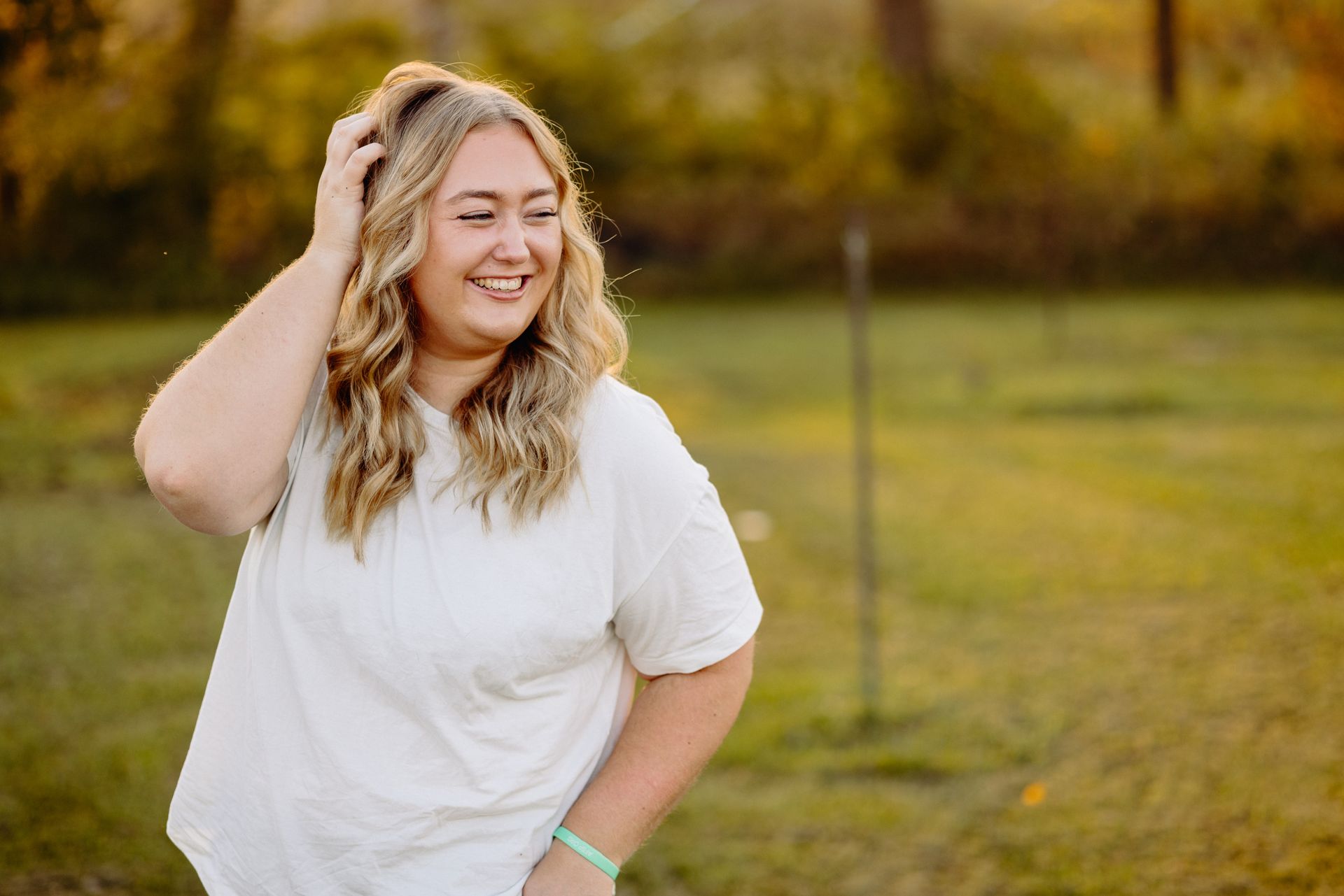 Woman with long, blonde hair smiles while touching her hair; wearing white shirt, standing in a grassy field.