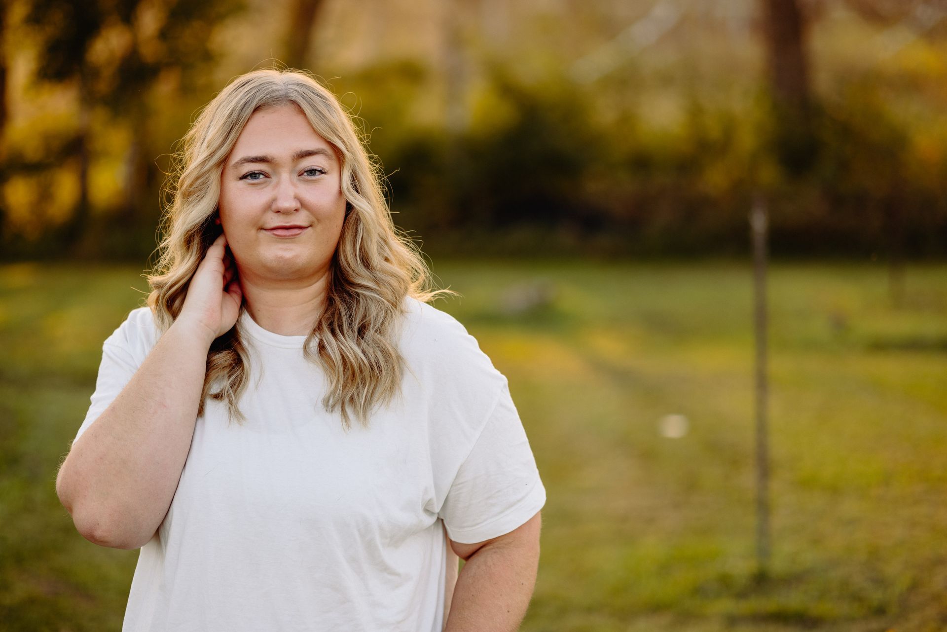 Woman who's a MN photographer with blonde hair, white shirt, hand on neck, smiling outside in golden light.
