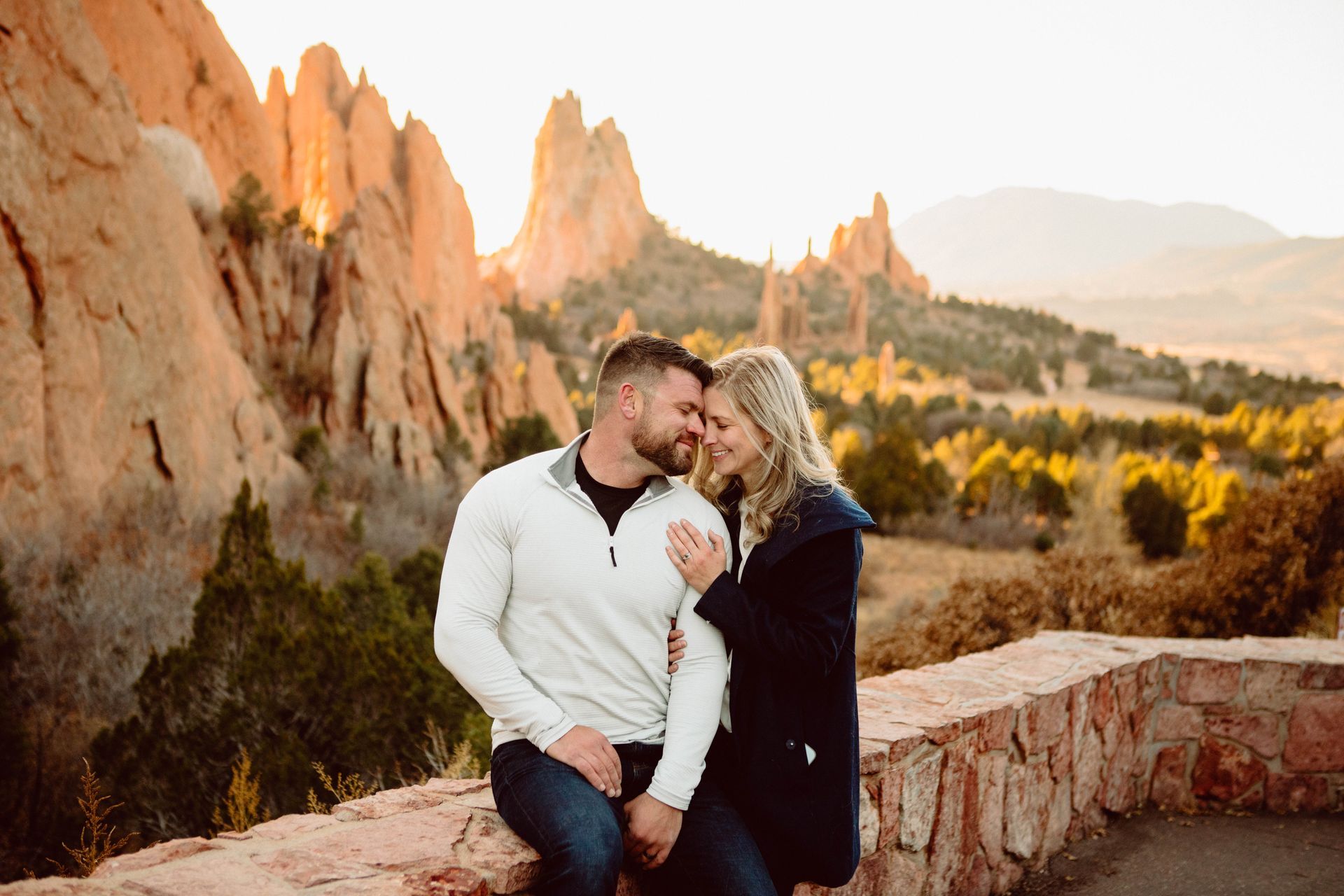 Couple embraces on a stone wall overlooking mountains at sunset.