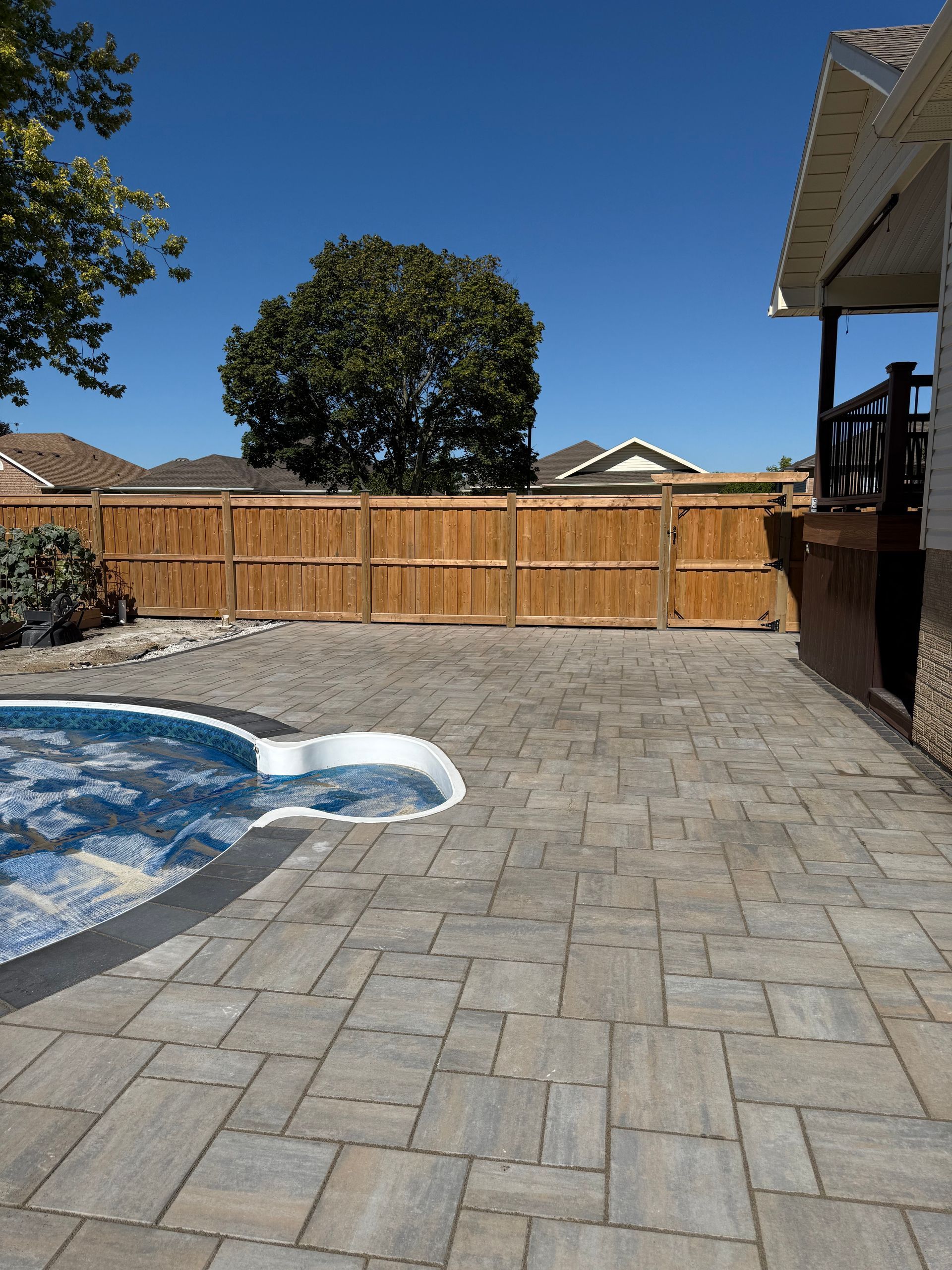 Backyard patio with a pool, stone pavers, wooden fence, and clear blue sky
