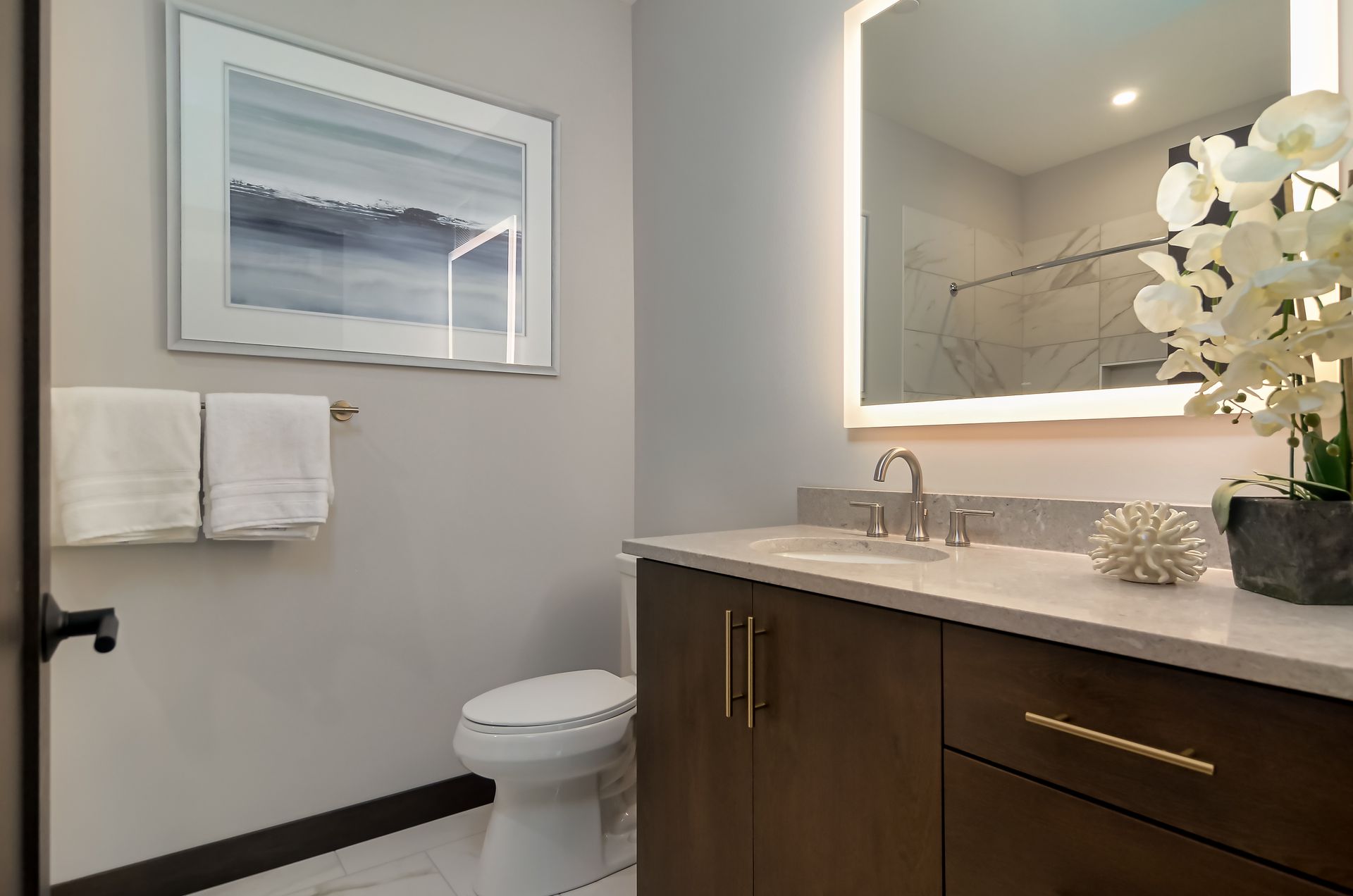 Bathroom with a large illuminated mirror, white countertop, dark wood vanity, toilet, and decorative orchid at Ventana Apartments in Wayzata, MN.