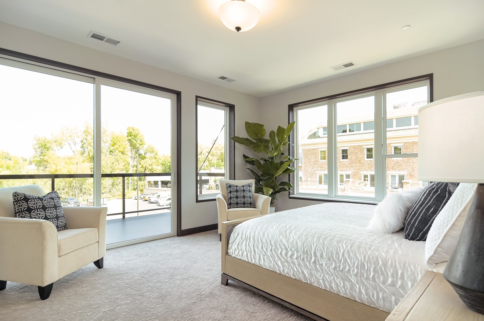 Bright bedroom with large windows, white bedding, gray carpet, and a sitting chair by the door Apartments in Wayzata, MN