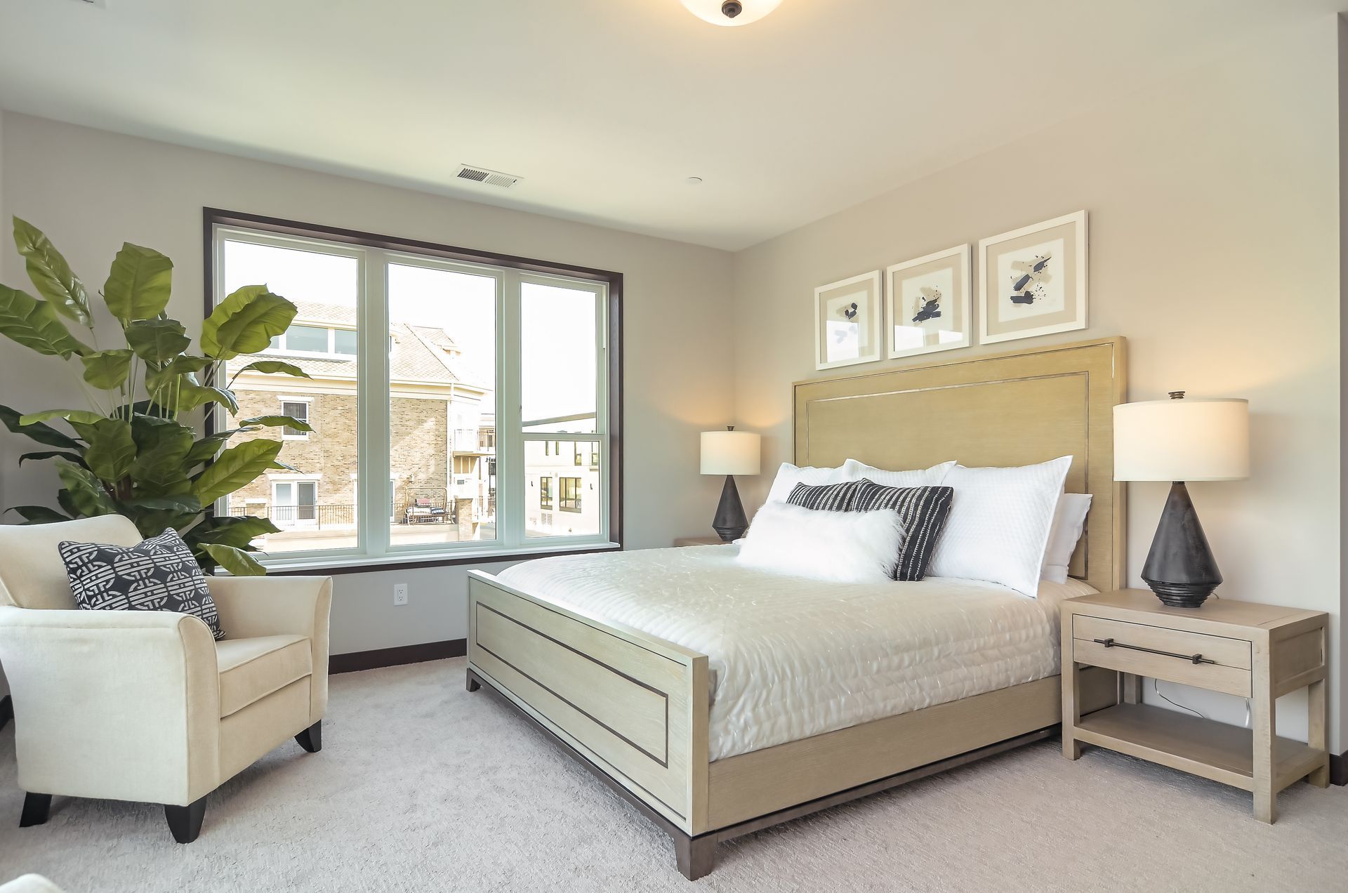 Bedroom with a large window, bed, armchair, nightstand, lamps, and framed art at Ventana Apartments in Wayzata, MN.