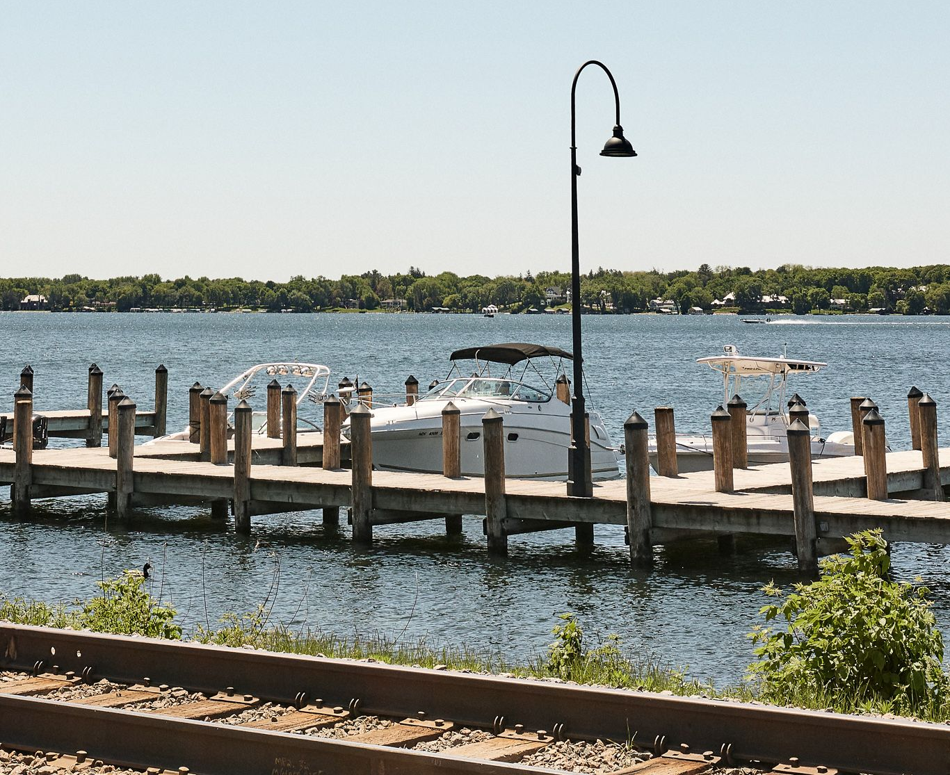 Wooden pier with parked boat, lamp post, and water, with trees on the horizon. Railroad tracks in foreground at Ventana Apartments in Wayzata, MN.