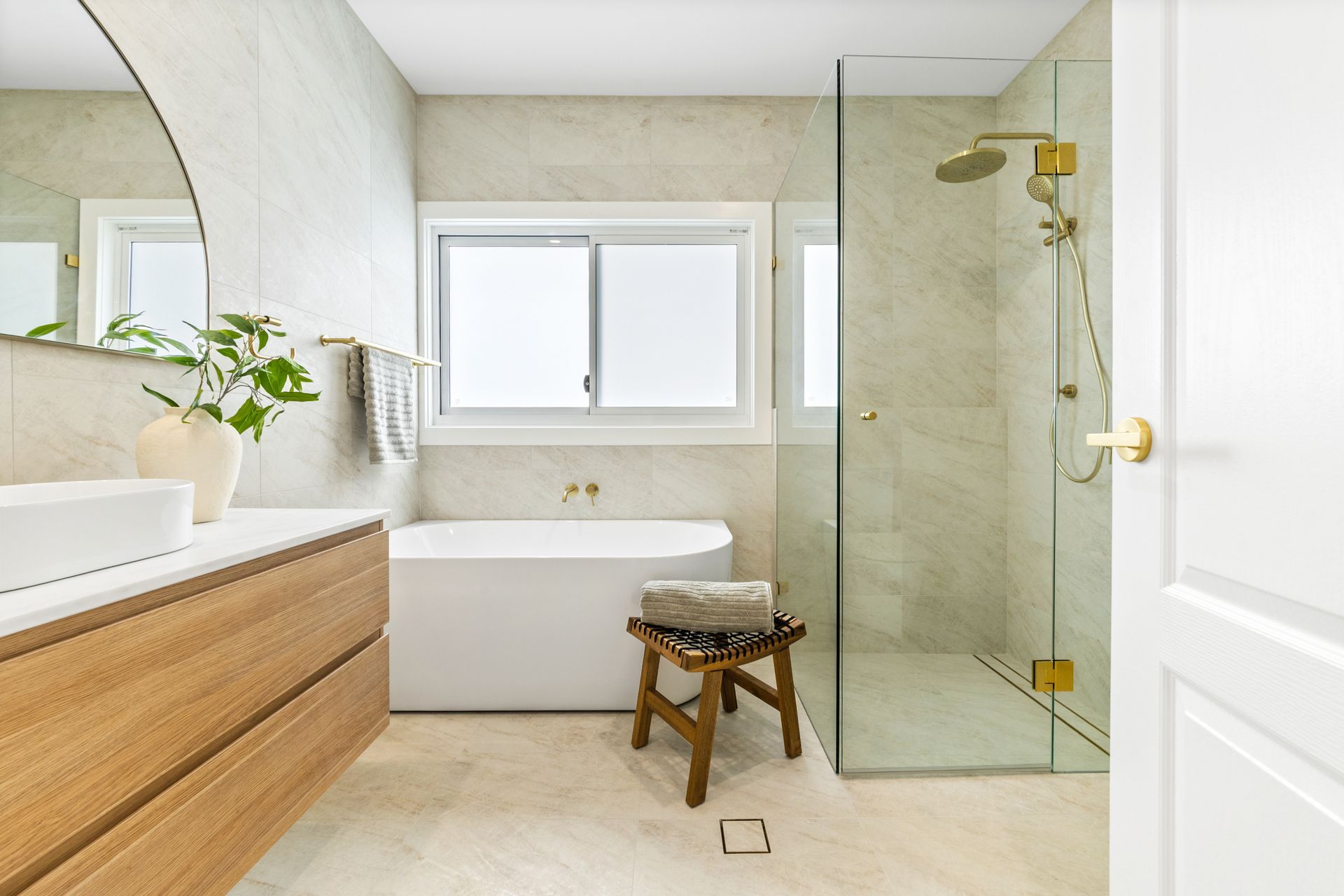 Bright modern bathroom with bathtub, wood vanity, glass shower, and small stool under a window — Designer Building Pty Ltd In Belmont, NSW