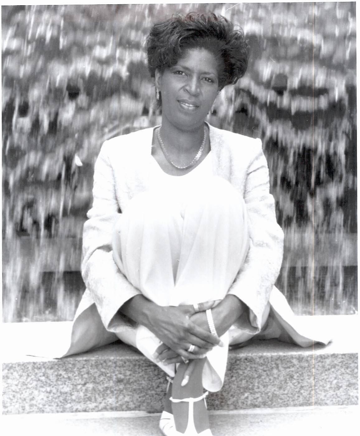 Woman sitting on a ledge in front of a waterfall, wearing a light suit and looking at the camera.