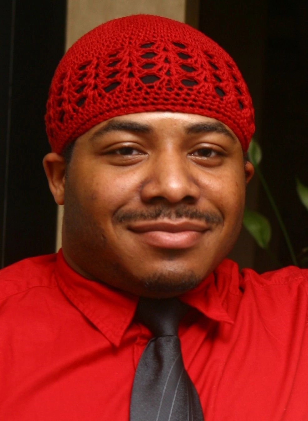 Man in red shirt and hat, smiling with a dark tie.