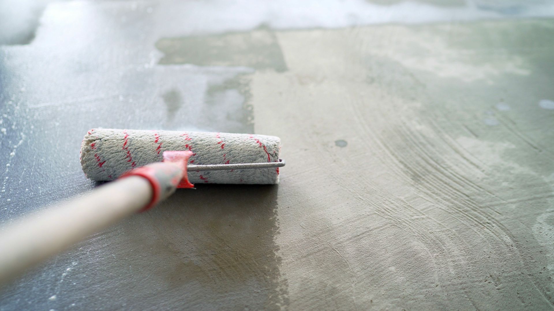 Paint roller applying a light-colored coating to a gray surface, possibly concrete.