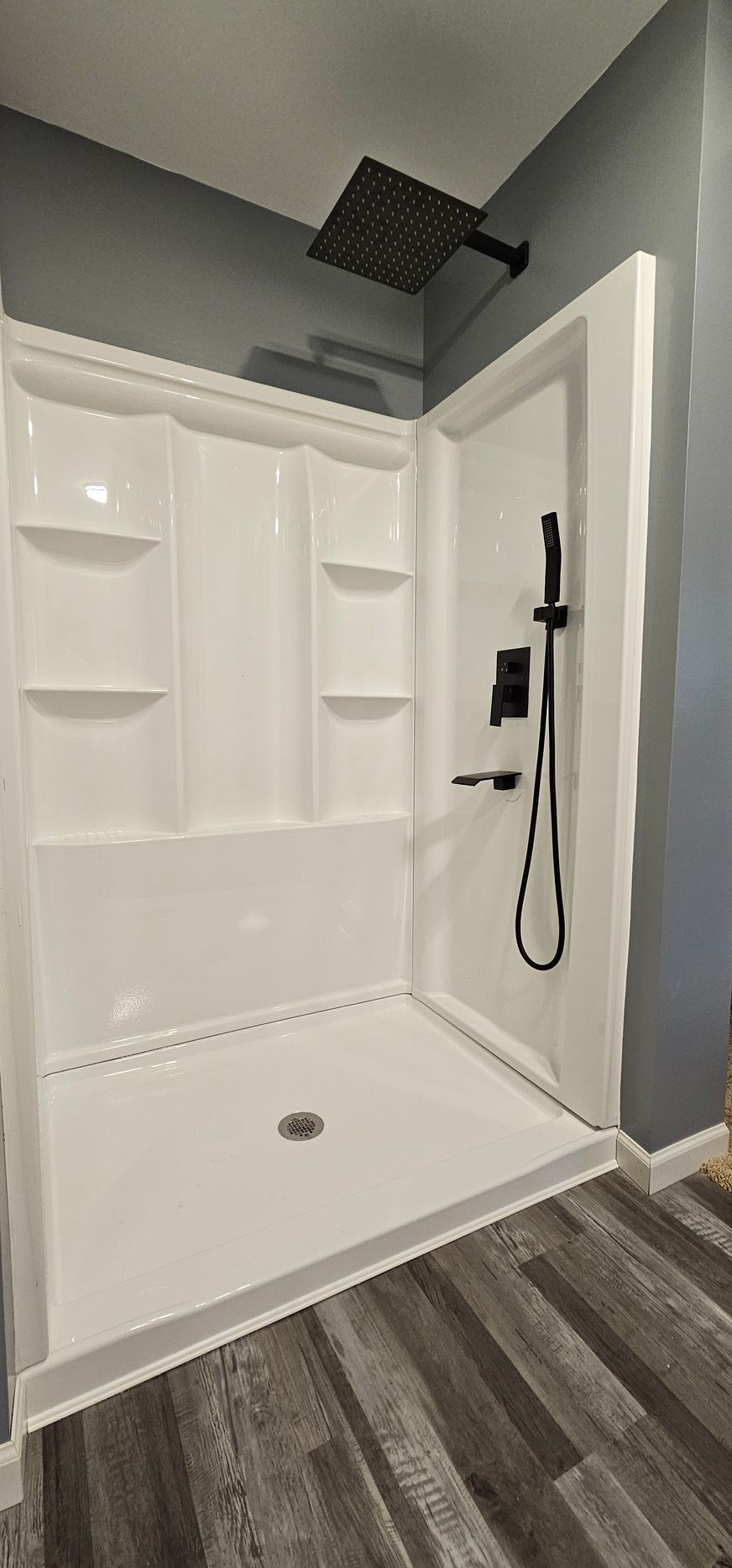 White walk-in shower with bench, black rainfall showerhead, and gray tile floor in a bathroom
