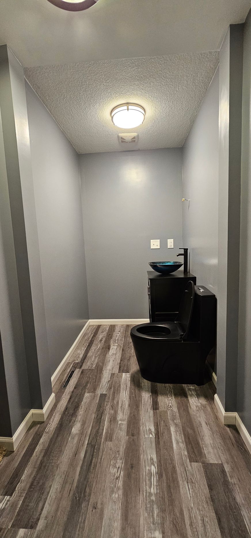Narrow gray hallway with wood-pattern floor and a black chair near a small sink or cabinet at the end.