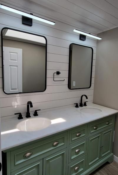 Bathroom vanity with twin sinks, black faucets, green cabinets, and two mirrors above white shiplap walls
