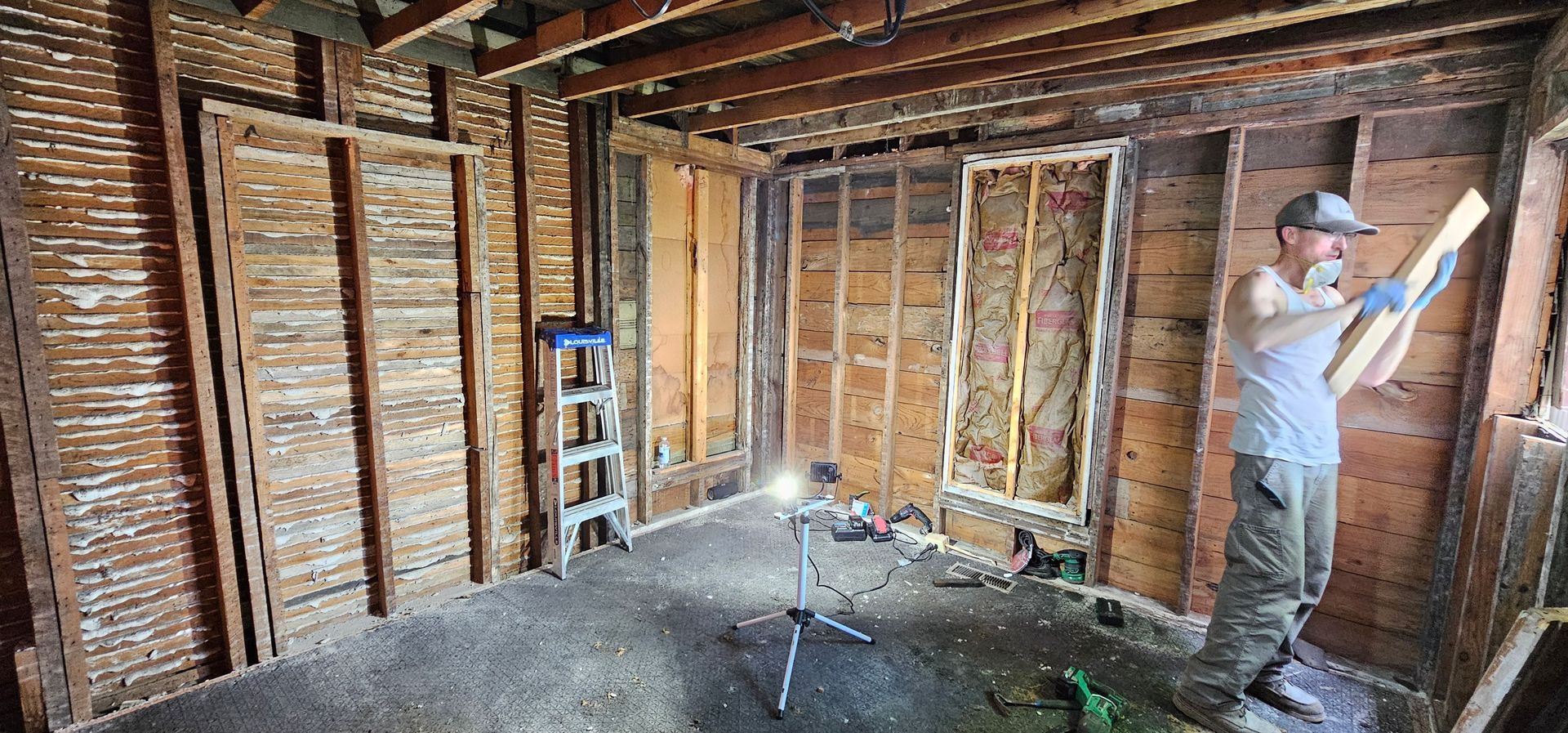 Person painting bare wooden walls inside a stripped-down room under renovation
