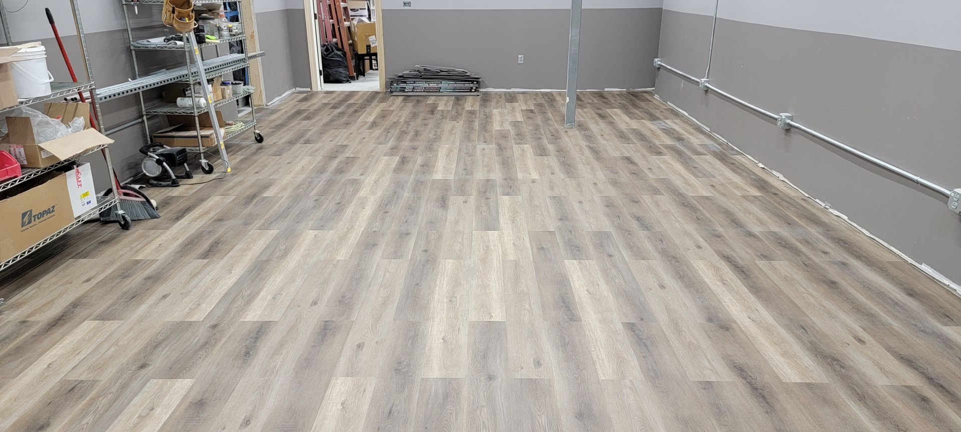 Empty warehouse with gray walls and light wood flooring, shelves and equipment along the left side.