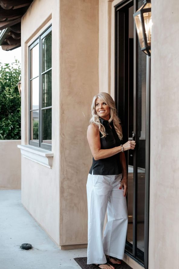 Woman standing in a doorway beside a beige stucco house, holding the door open and smiling