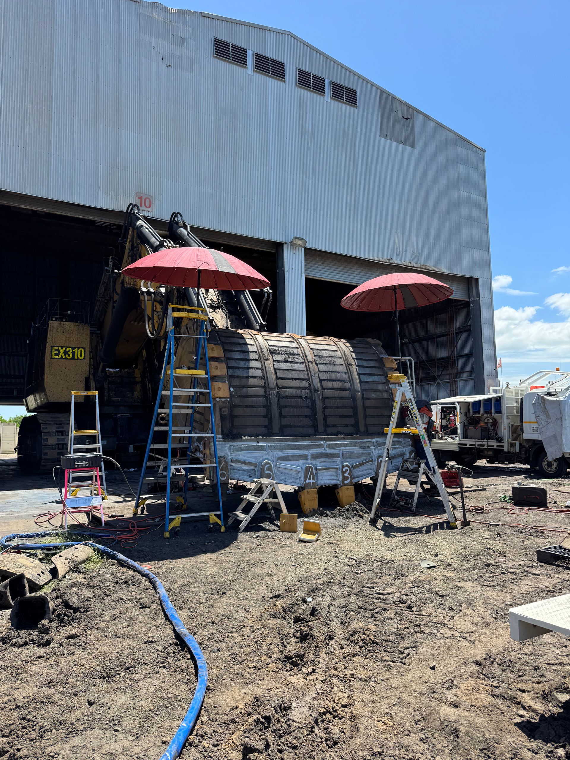 Workers on ladders under umbrellas outside a corrugated metal warehouse, with stacked materials and equipment — Resurge Diesel & Fabrication Pty Ltd In Balnagowan, QLD