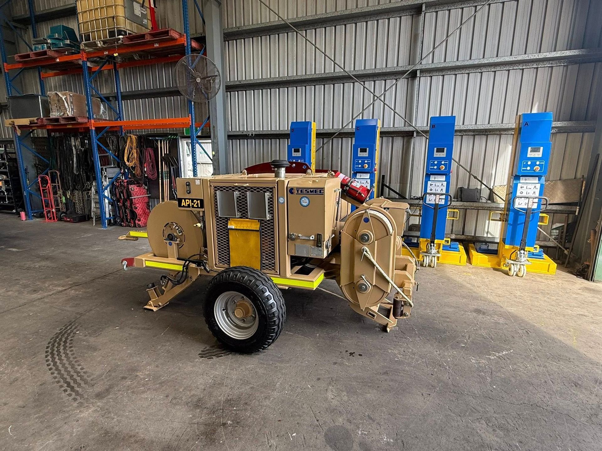 Tan agricultural machine in a warehouse beside blue hydraulic lifts and shelving