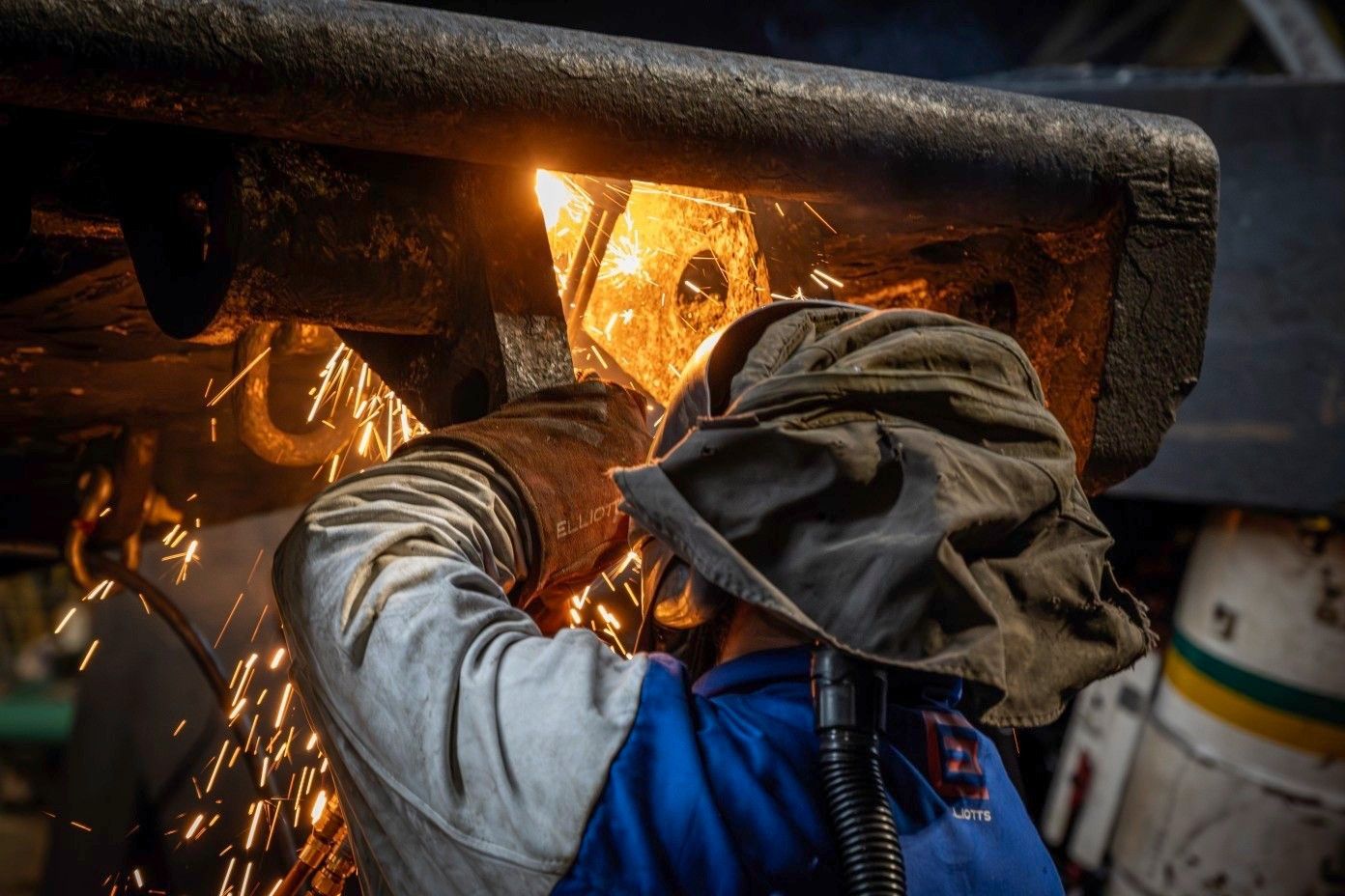 Worker using a welding torch under a metal frame, sparks flying in a dim workshop