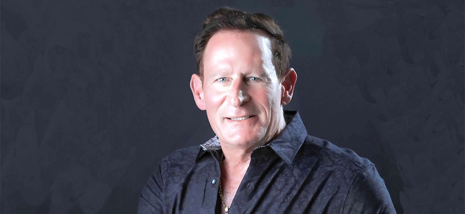 A man in a blue shirt is standing in front of a black background.