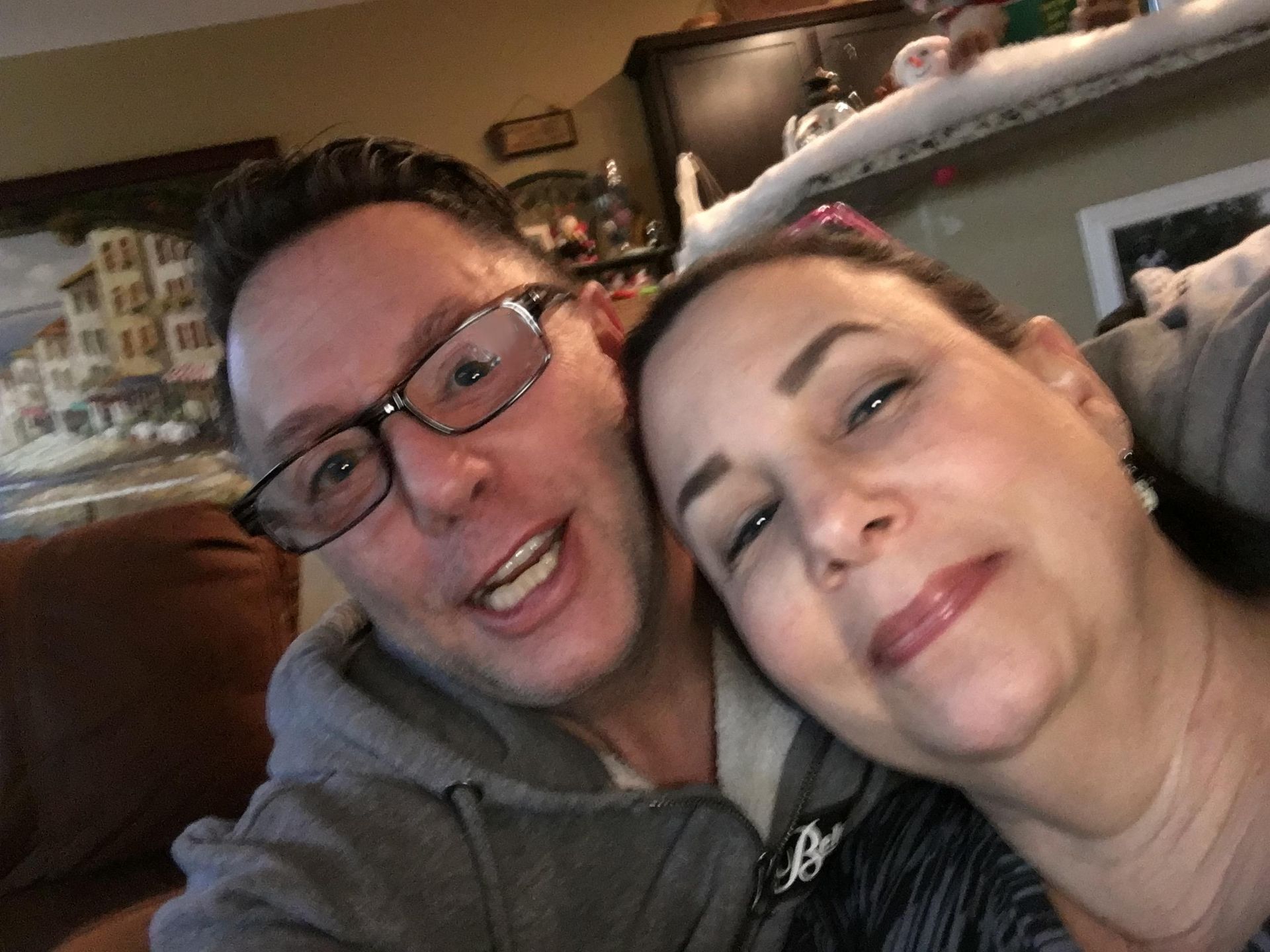 Two people smiling closely on a couch in a cozy living room, one wearing glasses and a gray hoodie.