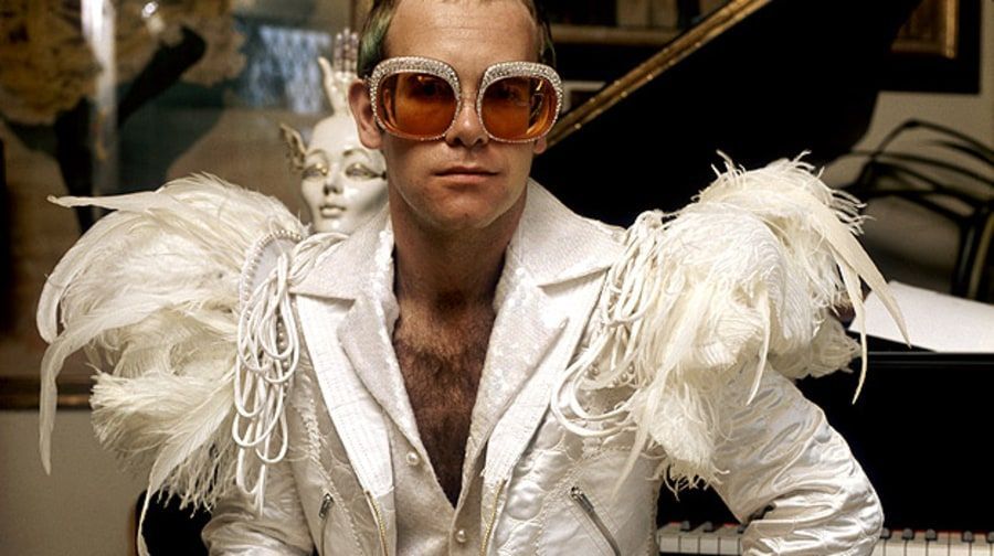 Man in oversized orange sunglasses and white feathered outfit, seated indoors on a black sofa