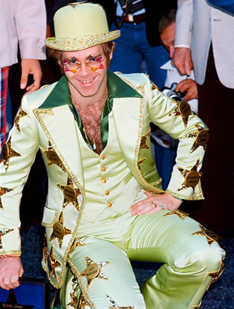 A man in a gold suit and hat poses seated, smiling, with star accents on his outfit.