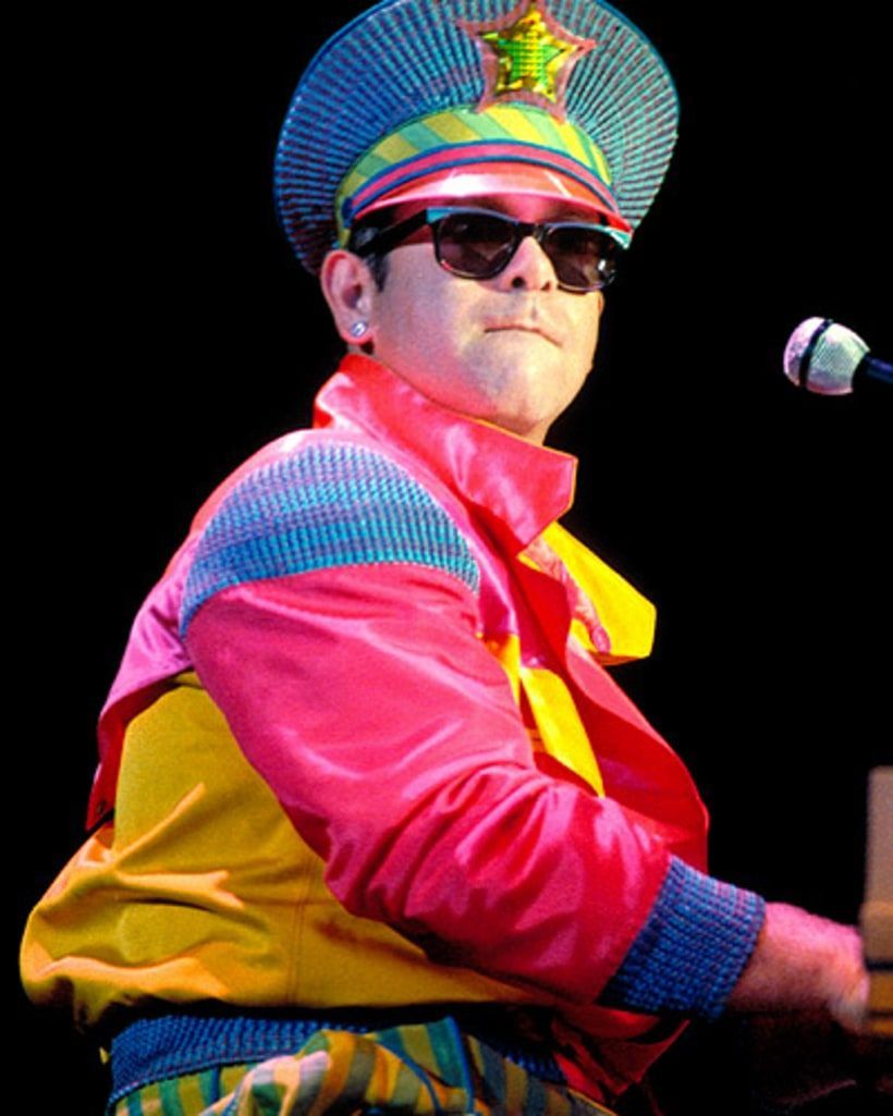 Person in colorful stage costume and sunglasses, seated at a microphone against a dark background.