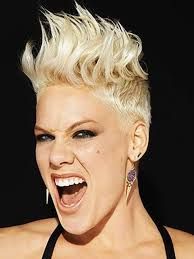 Close-up of a person with short platinum-blond spiked hair, mouth open in a fierce expression on a dark background