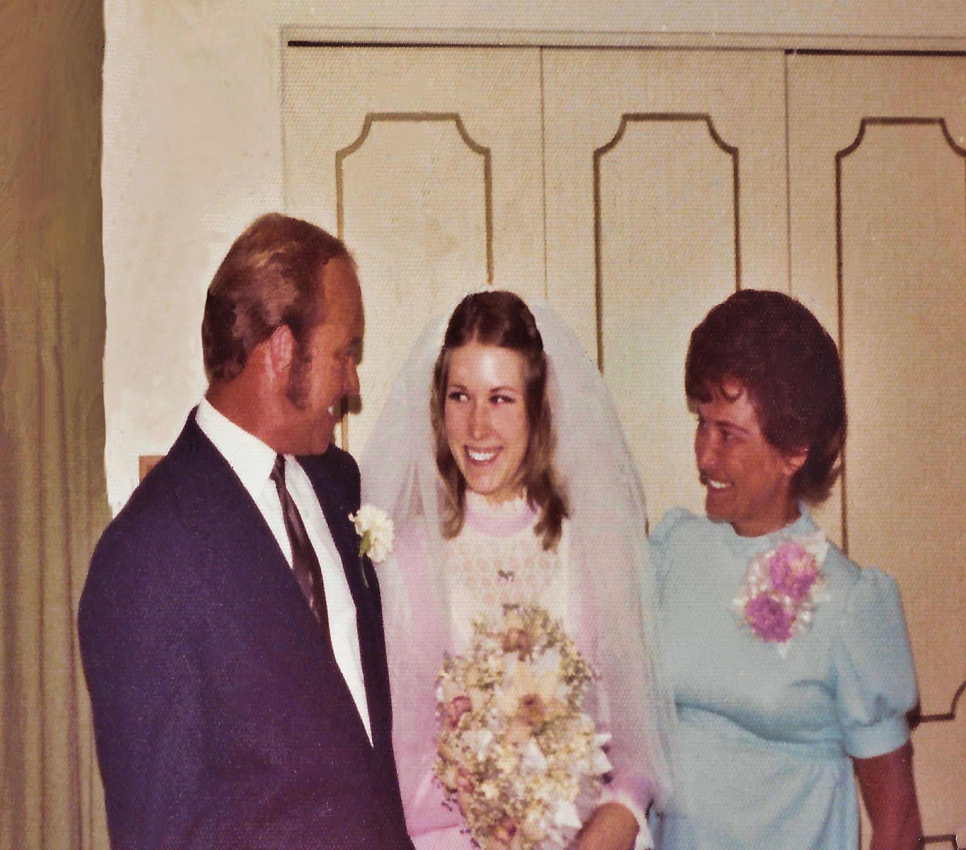 Three people standing indoors, with a bride in a veil holding a bouquet between two guests.