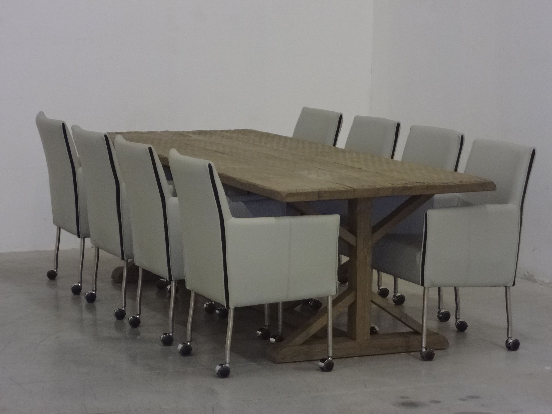 Een rechthoekige houten eettafel met een kruisvormig onderstel, omringd door acht lichtgrijze gestoffeerde stoelen op wielen.