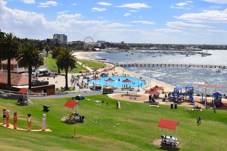 Beachside park with Ferris wheel, pool, playground, pier, and harbor under a sunny sky.