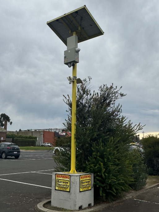 Yellow pole with solar panel, camera, and warning signs in a parking lot.