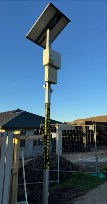 Solar-powered pole with equipment, likely for monitoring, with a measuring scale. New construction in the background.