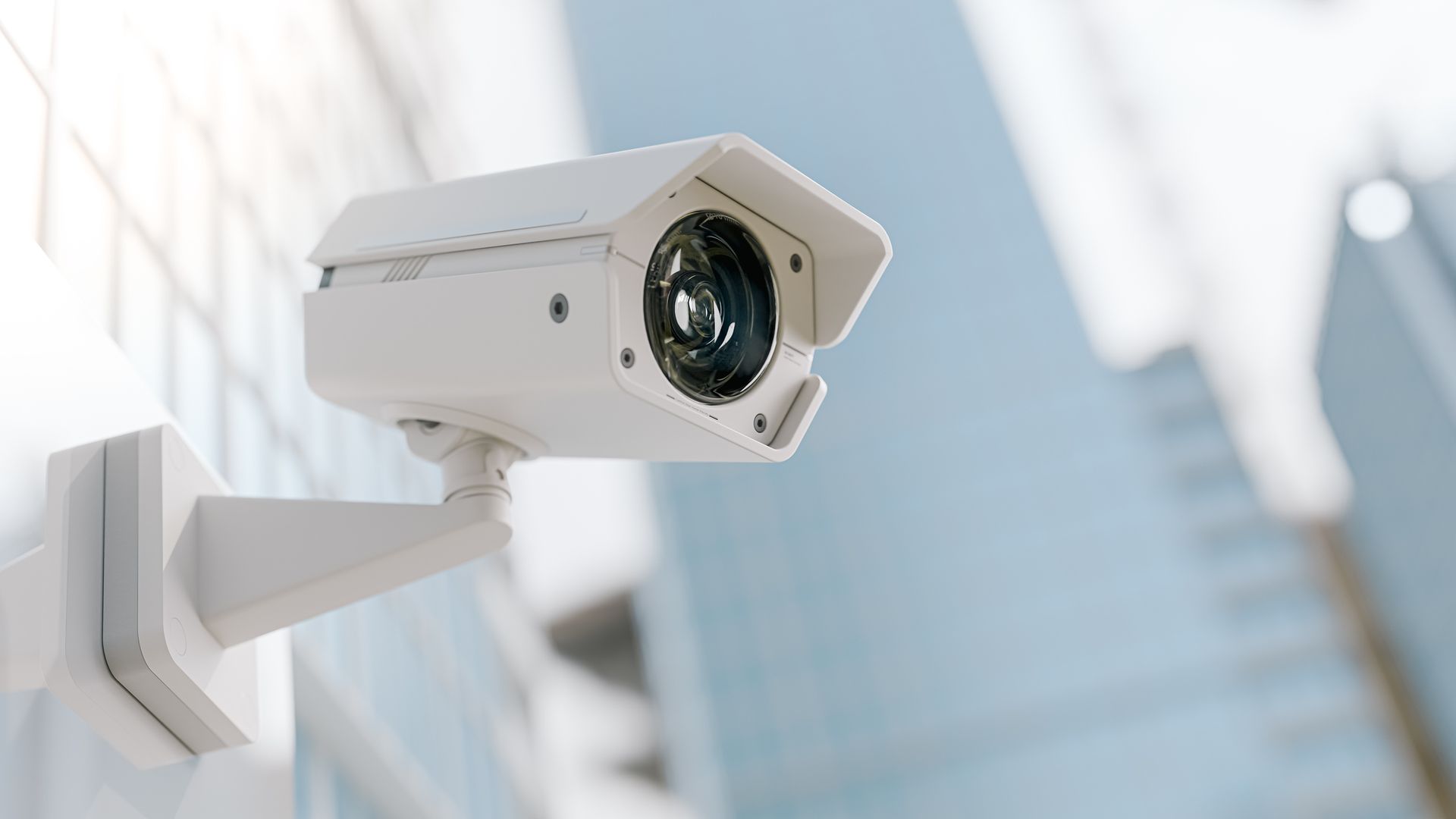 White CCTV security camera mounted on a building wall with blurred city skyscrapers in the background.