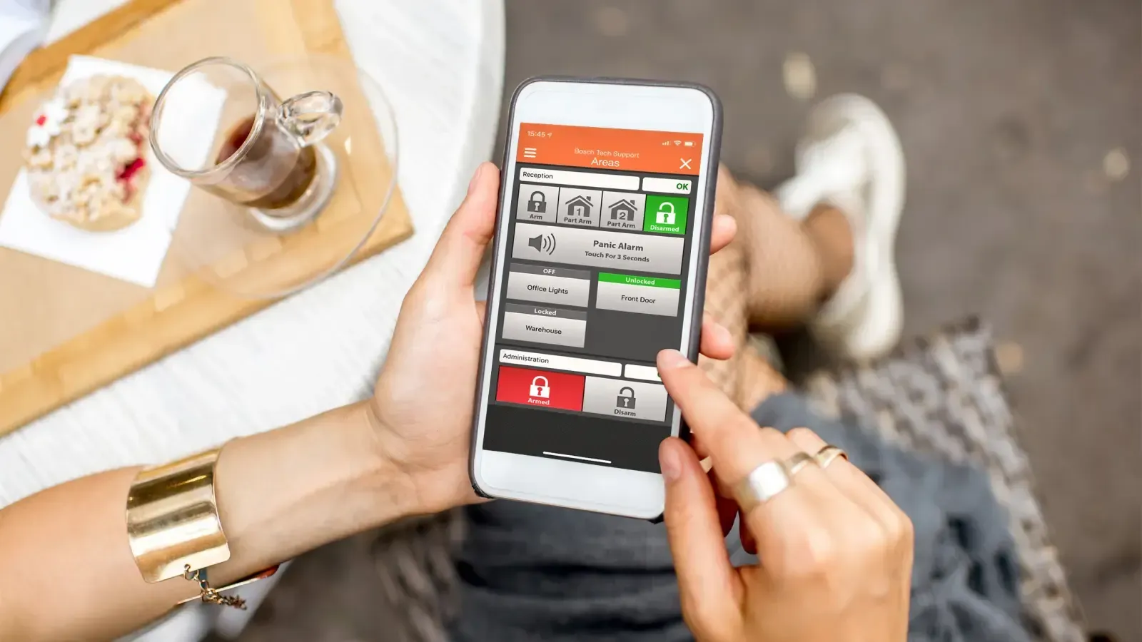 Person using a phone with a smart home app. Buttons for security functions displayed, outdoors, coffee cup visible.