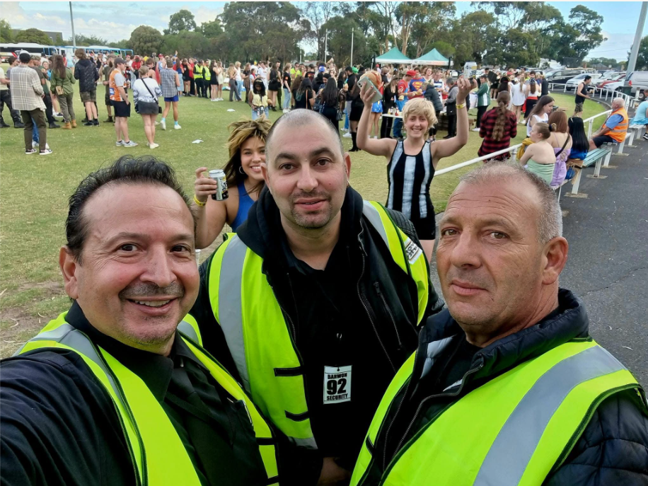 Three people in safety vests take a selfie at an outdoor event, crowd in background.